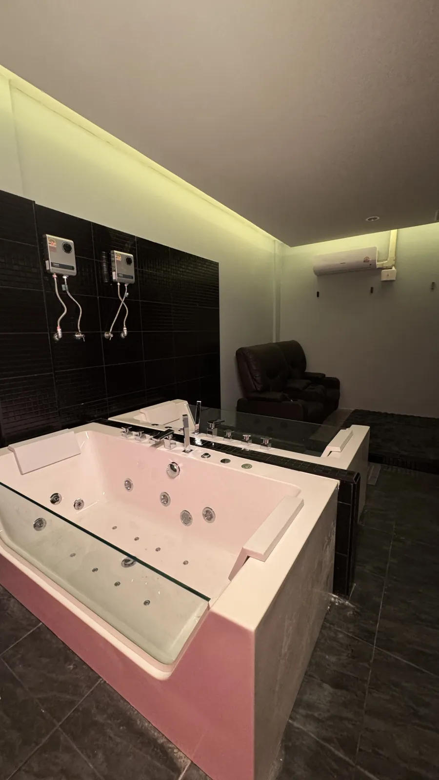 VIP jacuzzi suite at Heal, Bangkok — a glass-sided whirlpool tub beside a black-tiled wall and a private leather recliner, dim light.