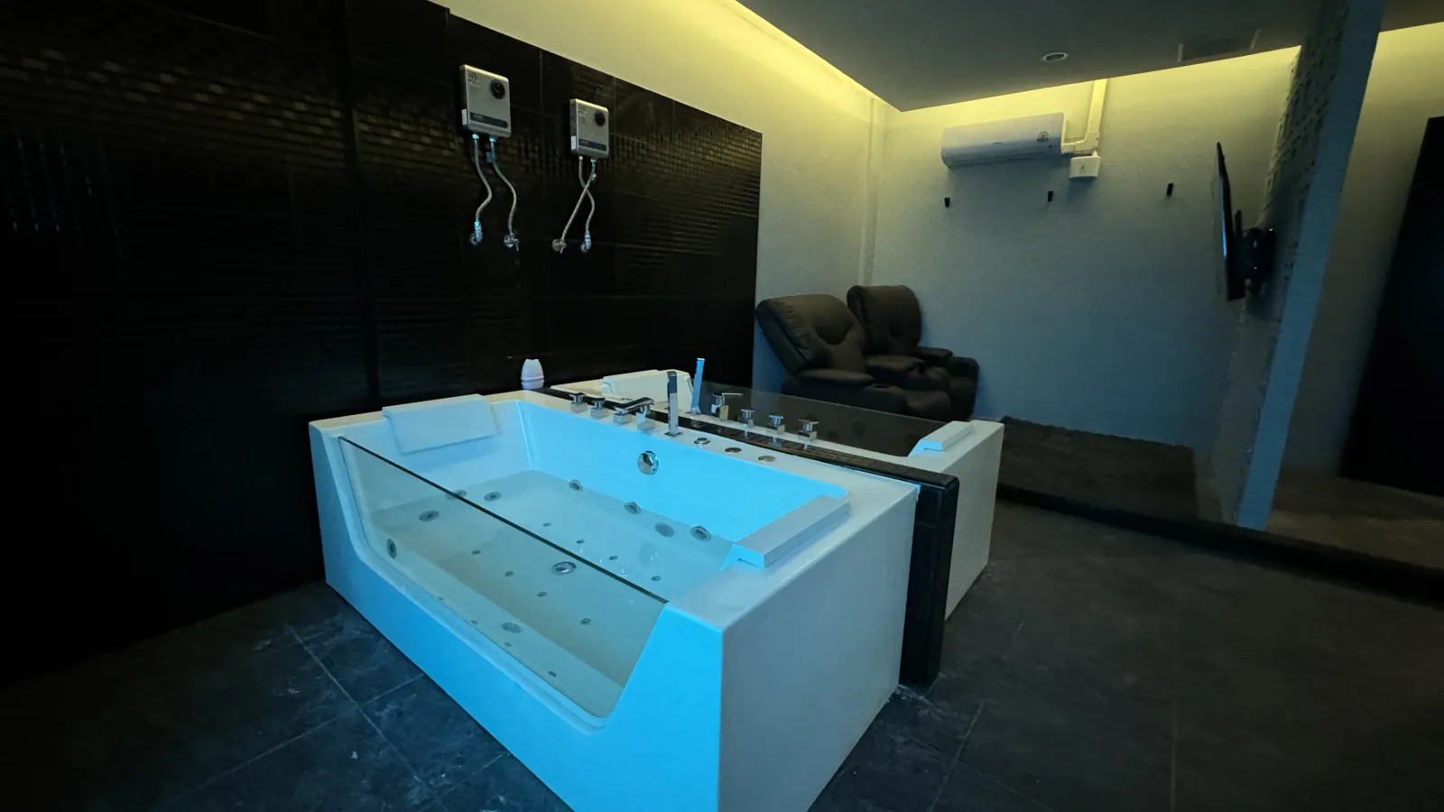 Jacuzzi suite at Heal nuru massage studio, Bangkok — a large glass-sided whirlpool tub with private lounge recliners and a wall TV, dim modern setting.